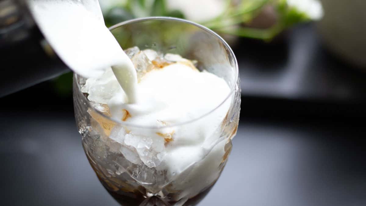 A pitcher pouring frothed milk over an iced coffee in a crystal glass