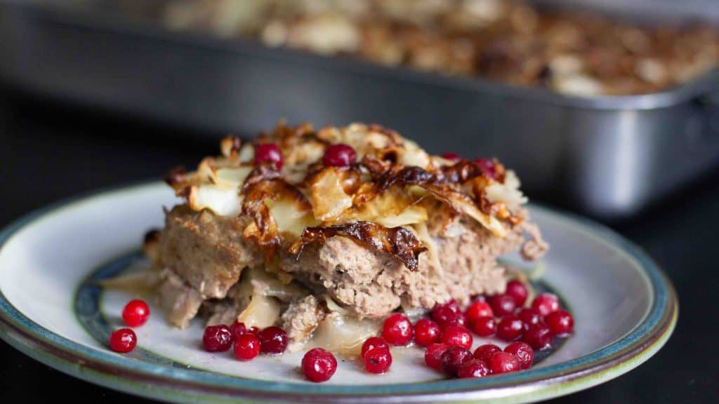 side view of Swedish Cabbage Meatloaf - Kålpudding served on a ceramic plate with rawLingonbery Sauce. There is a dish of kålpudding in the background
