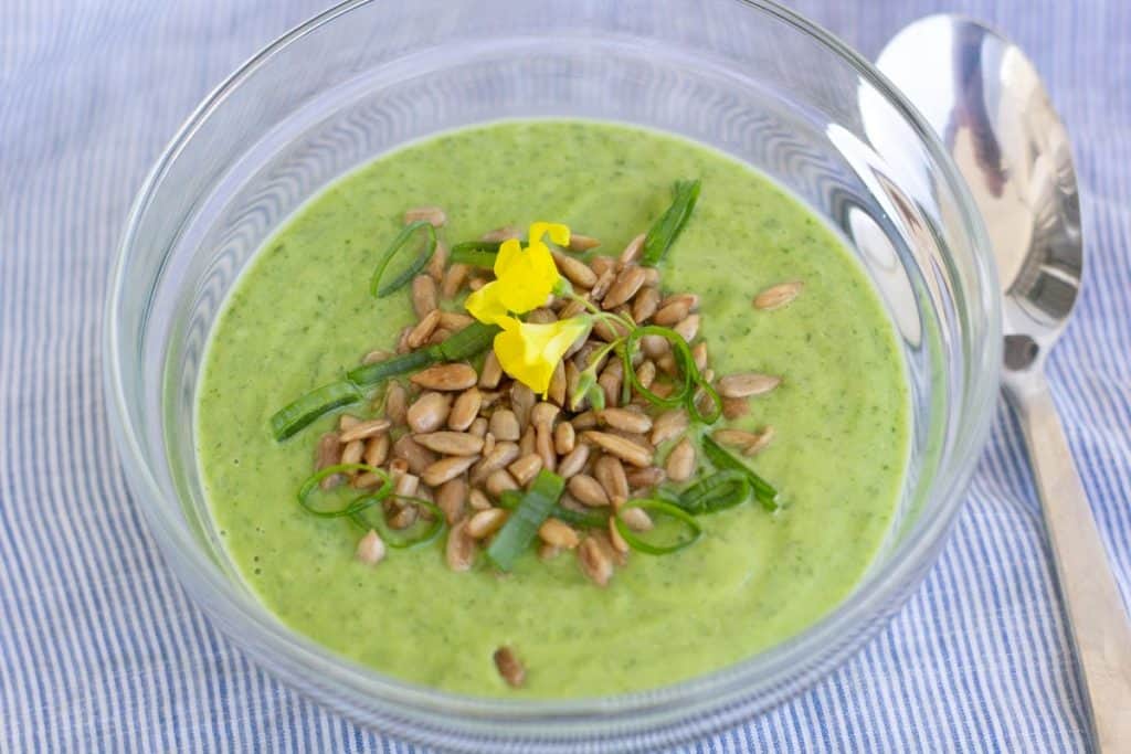 Chilled sorrel soup served in a glass bowl with a spoon beside. the sou is garnished with salted sunflower seeds, finely chopped spring onion and wood sorrel blossoms