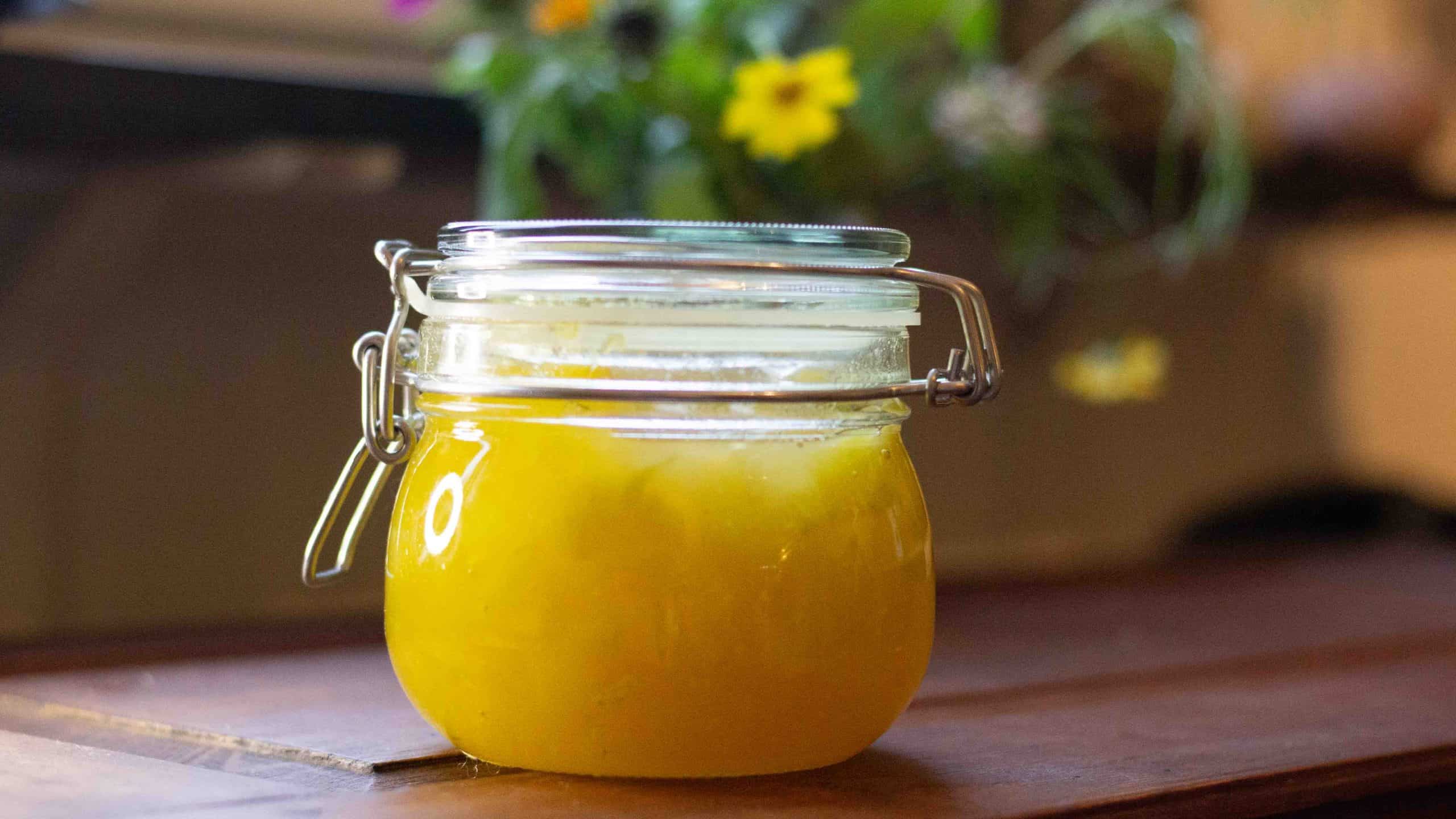 Bright yellow marrow jam in a glass jar on a wooden table. There are garden flowers in the background