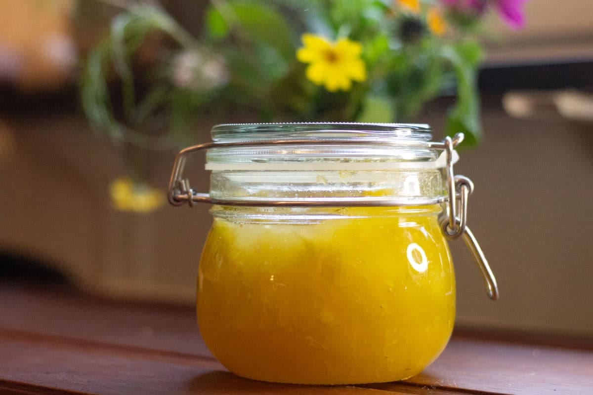 a glas jar of bright yellow marrow jam on a wooden table with yellow adn pink flowers in the background