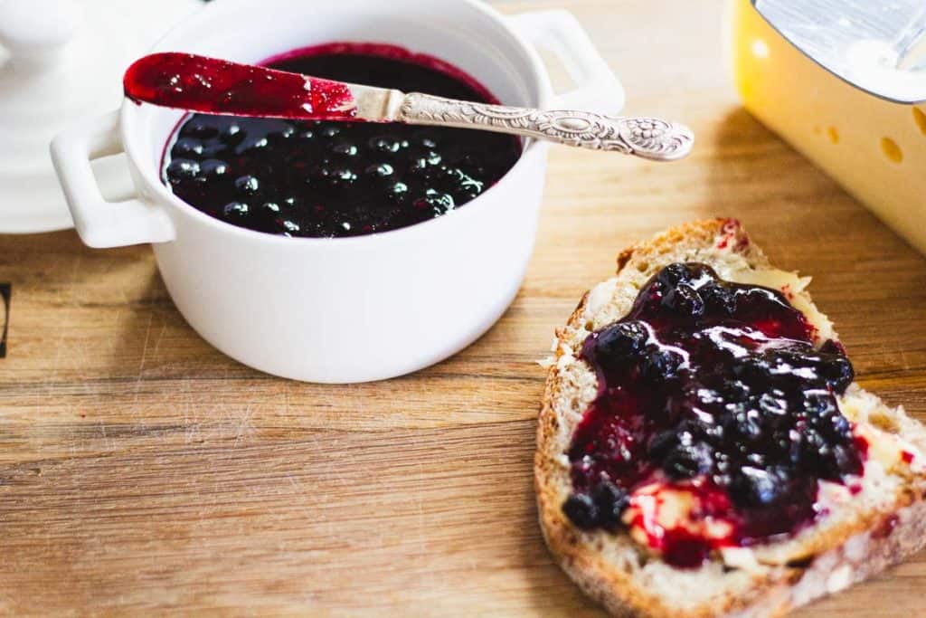 a silver butter knife resting on a pot of queens jam. Beside it a slice of buttered toast with jam spread on it and a block of cheese