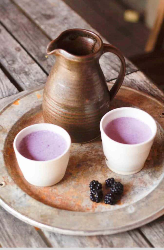two ceramic cups of blackberry milk on a tray with some wild blackberries and a pottery jug