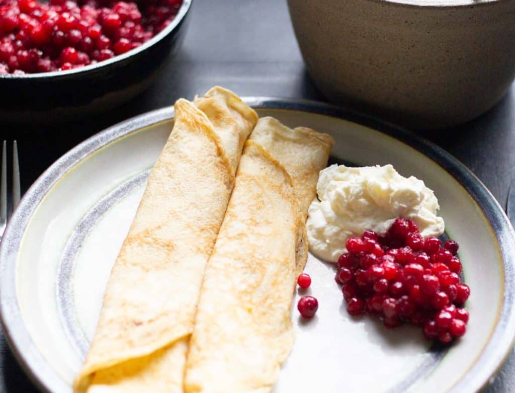 2 Swedish pancakes served with whipped cream and lingonberry sauce on a ceramic plate
