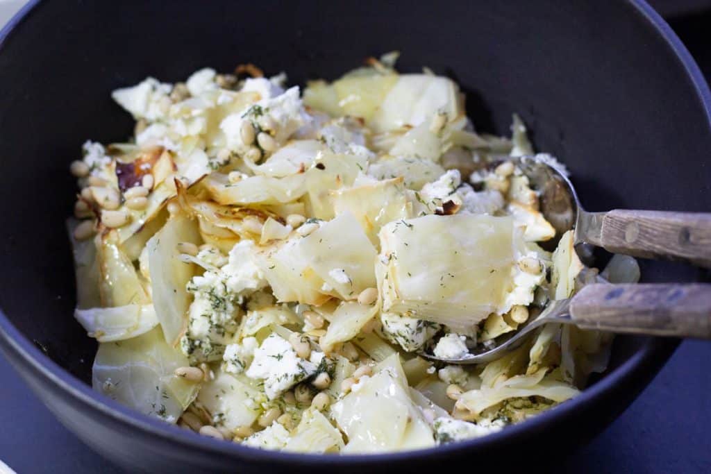 s large black ceramic bowl filled with roasted cabbge salad with wooden salad spoons