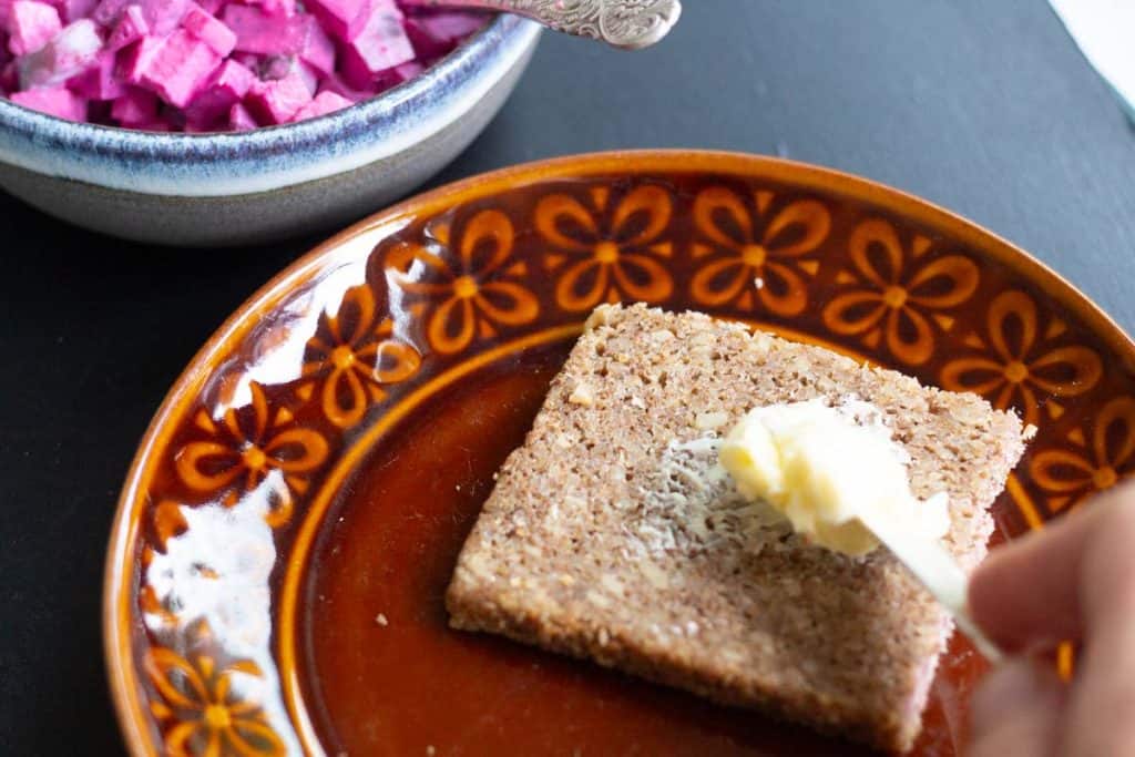 A knife spreading butter on Swedish rye bread that is on a plate beside a dish of Swedish beetroot and apple salad