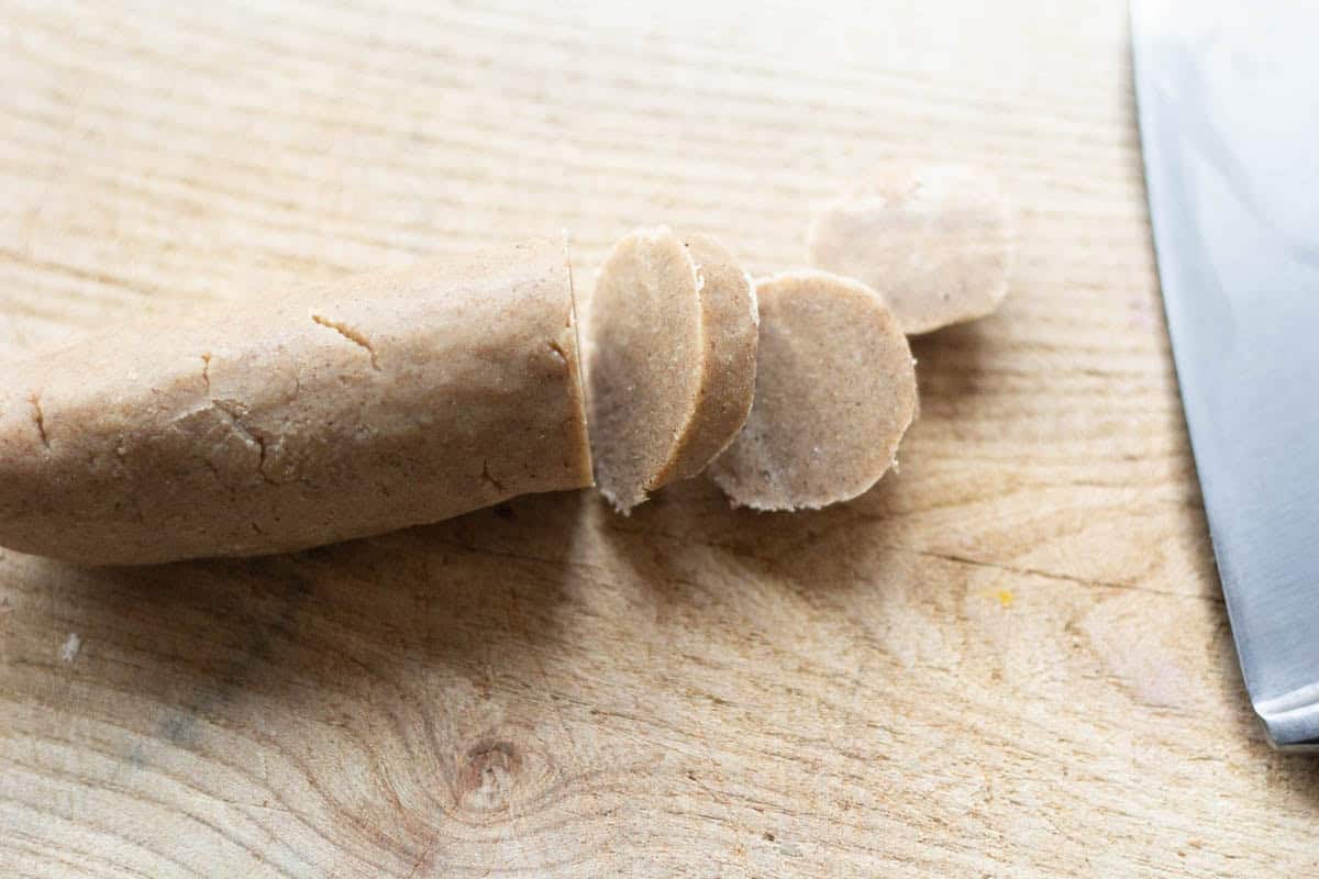 a sharp knife beside gingerbread shortbread cookie dough that is sliced into rounds on a wooden chopping board