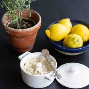 Bone Mrrow Butter in a white ramekin dish with a silver knife. The lids of the dish is beise it together with a blue ceramic bowl filled with fresh lemons and a pot of fresh rosemary