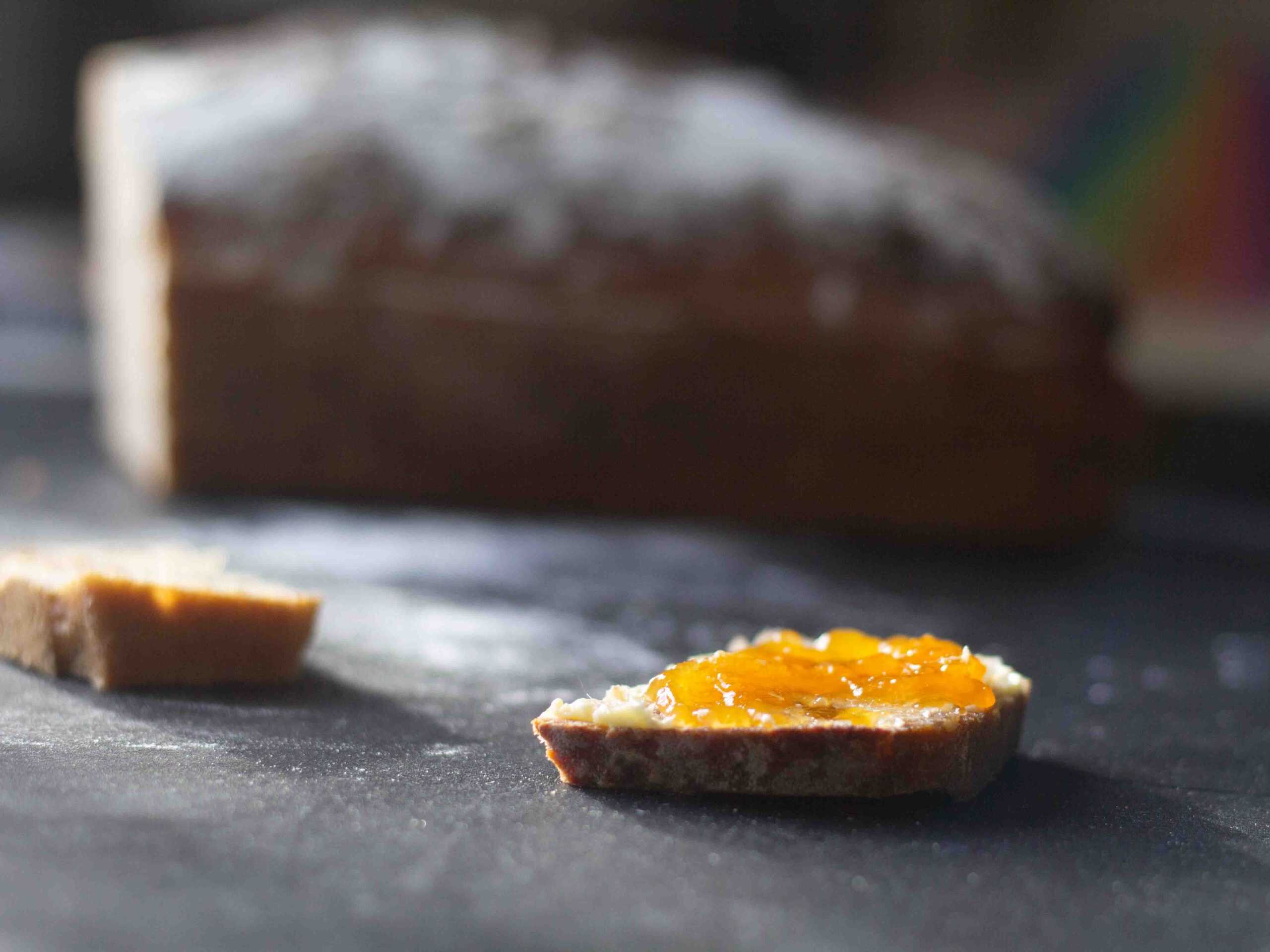 side view of a slice of sourdough bread with cloudberry jam on top. there is a loaf od bread in the background