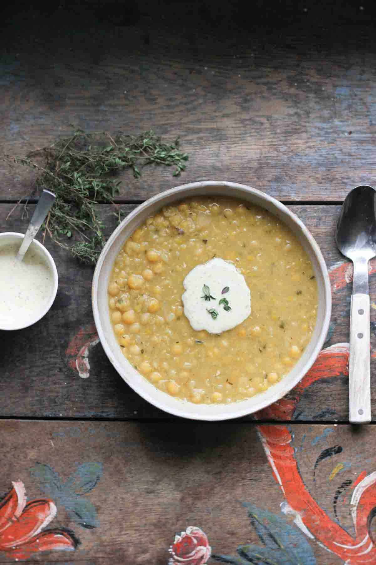 ärtsoppa or Swedish pea soup served in a white cermaic bowl and topped with wholegrain mustard sauce and some freh thyme. The bowl is on a painted wooden table