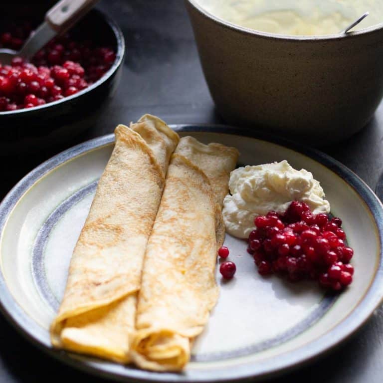 Two Swedish pancakes rolled up on a ceramic plate beside lingonberry jam and whipped cream