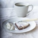 A slice of swedish sticky chocolate cake beside a dollop of whipped cream on a white plate with a fork. There is a cup of coffee in the background