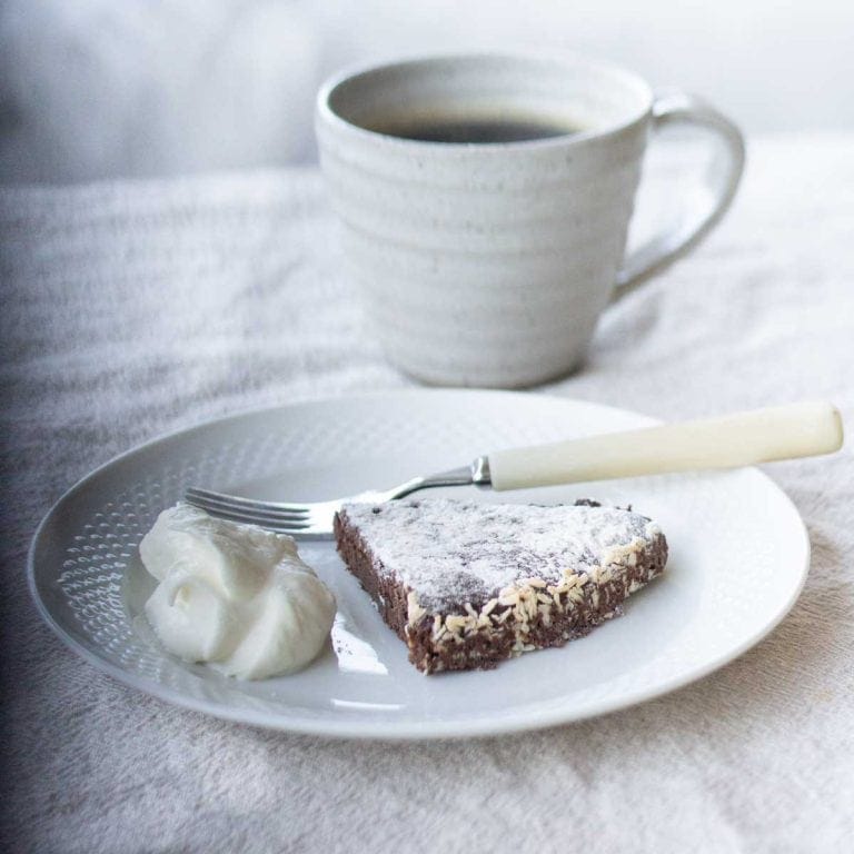 A slice of swedish sticky chocolate cake beside a dollop of whipped cream on a white plate with a fork. There is a cup of coffee in the background