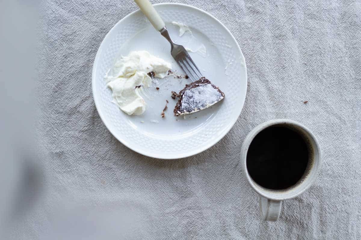 overhead image of a white plate with a half eaten slice of kladdkaka and cream served beside a ceramic cup of black coffee